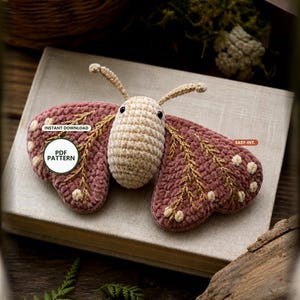 Puede incluir: Un peluche de polilla tejido a ganchillo con cuerpo color crema y alas rosa polvoriento con patrones de ramas doradas bordadas y pequeños puntos crema. La polilla tiene dos antenas y ojos negros. El texto dice: INSTANT DOWNLOAD, PDF PATTERN, EASY-INT.