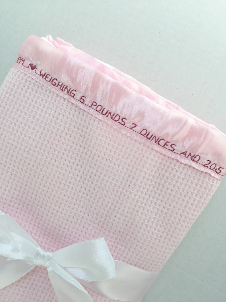 May include: A pink waffle weave baby blanket with a white satin ribbon tied in a bow. The blanket has a pink satin border with the embroidered text "Weighing 6 pounds 7 ounces and 2015."
