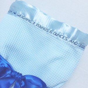 May include: A light blue waffle weave baby blanket with a satin trim and a blue satin ribbon tied in a bow. The trim has the text "Weighing 7 pounds 6 ounces and 20.5"