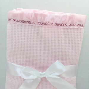 May include: A pink waffle weave baby blanket with a white satin ribbon tied in a bow. The blanket has a pink satin border with the text "BM &hearts; WEIGHING 6 POUNDS 7 OUNCES AND 20.5"