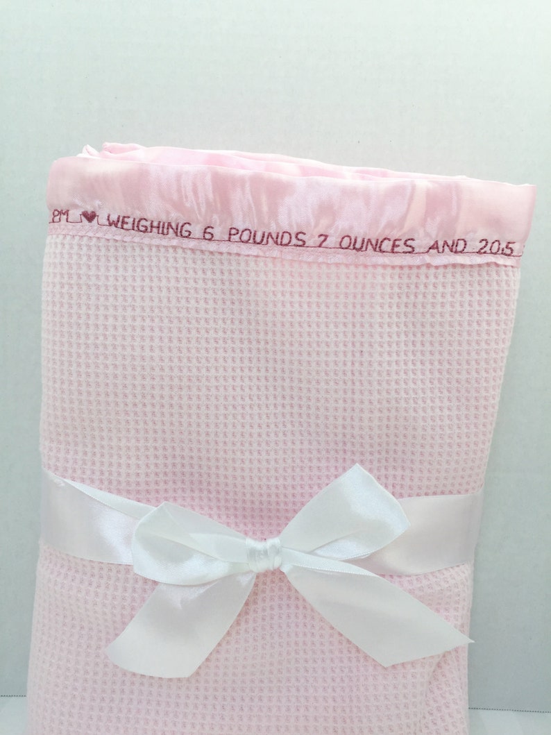 May include: A pink waffle weave baby blanket with a white satin ribbon tied in a bow. The blanket has a pink satin border with the text "BM &hearts; WEIGHING 6 POUNDS 7 OUNCES AND 20.5"