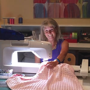 May include: A woman is using a sewing machine to sew a pink quilted fabric. The fabric is on a green cutting mat. The sewing machine has a bright light shining on the fabric.