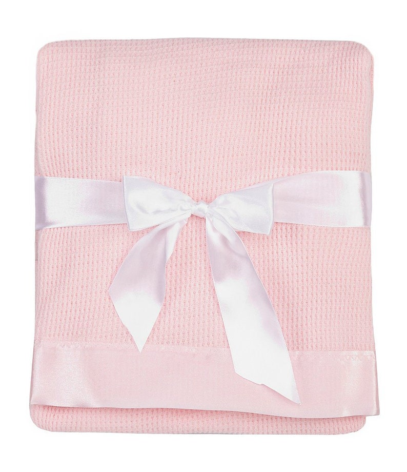 May include: A pink waffle weave baby blanket with a white satin ribbon tied in a bow. The blanket has a satin trim around the edges.