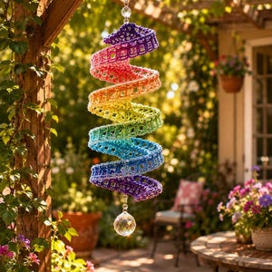May include: A spiral-shaped hanging decoration featuring a gradient of crocheted yarn in colors ranging from purple to pink, orange, yellow, green, and blue. The structure is suspended by a silver chain and accented with clear crystal beads.