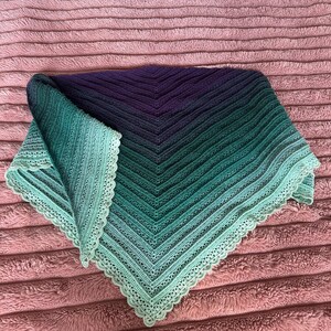 May include: A triangular crochet shawl featuring a gradient color transition from deep purple at the center to dark teal, transitioning into a lighter mint green at the edges. The shawl has a textured ribbed pattern and a decorative scalloped border.