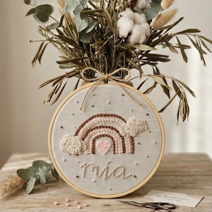 Könnte beinhalten: Ein runder Stickrahmen aus Holz mit beigefarbenem Stoff, der ein handgesticktes Design aus einem braun-rosa Regenbogen, einem rosa Herz und flauschigen weißen Wolken zeigt. Der Name "mia" ist darunter in Schreibschrift eingestickt.