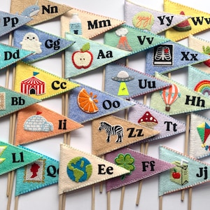 Op de afbeelding: Een set vlaggetjes van vilt met letters van het alfabet en verschillende illustraties. De vlaggetjes zijn in verschillende kleuren, waaronder blauw, groen, geel, oranje, roze en rood. De illustraties omvatten een spook, een zebra, een tent, een sapdoos, een appel, een vulkaan, een luchtballon, een UFO, een paddenstoel, een doos met kleurpotloden, een aardbol, een hart, een ster, een regenboog, een sinaasappel, een iglo en een schedel.