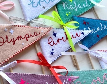 May include: A collection of felt pennants with names written in white script. The pennants are decorated with white stars and are attached to wooden sticks with colorful ribbons.