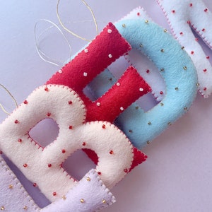 May include: A set of felt letter ornaments in the colors red, white, blue, and purple. Each letter is decorated with small beads.