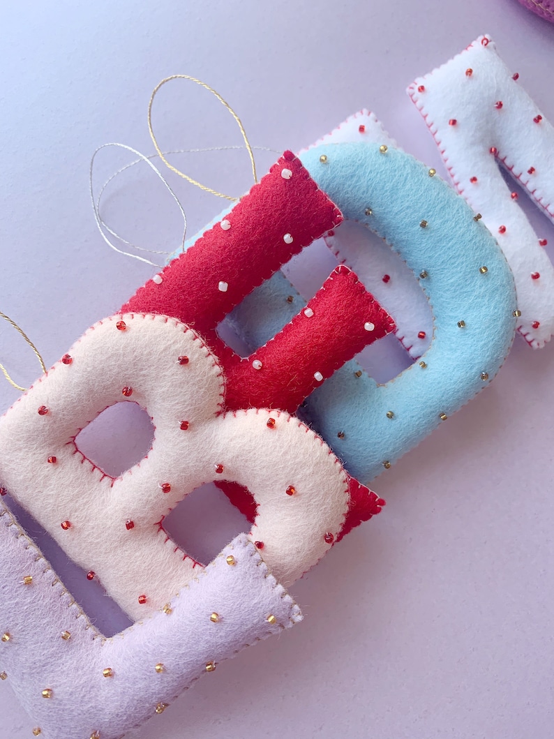 May include: A set of felt letter ornaments in the colors red, white, blue, and purple. Each letter is decorated with small beads.