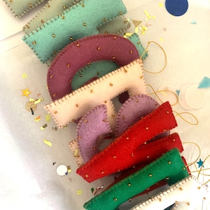 May include: A set of felt letters with gold bead accents, spelling out the word "MADE". The letters are in various colors, including green, pink, purple, red, and white.