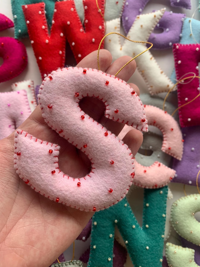 May include: A pink felt letter S with red stitching and white beads. The letter is hanging from a gold string and is surrounded by other felt letters.