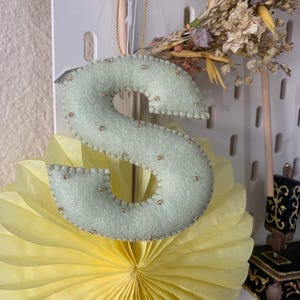 May include: A light green felt letter S with gold beads sewn around the edges. The letter is hanging from a gold string and is in front of a yellow paper fan.