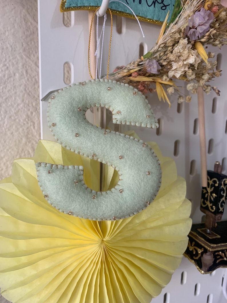 May include: A light green felt letter S with gold beads sewn around the edges. The letter is hanging from a gold string and is in front of a yellow paper fan.