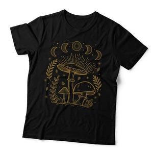 May include: A black short-sleeved t-shirt featuring a gold-coloured line art graphic on the front. The design displays three mushrooms surrounded by foliage, crystals, and a series of moon phases above, all rendered in thin, detailed golden lines.
