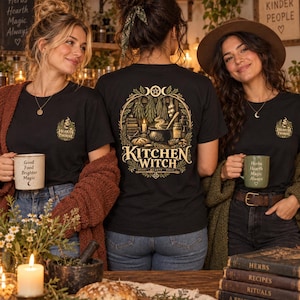 Op de afbeelding: Een zwart t-shirt met een afbeelding op de achterkant met de tekst "KITCHEN WITCH" en "HERBS HEARTH MAGIC" onder een illustratie van een ketel, kruiden en kaarsen. Op de linkerborst staat een klein logo met de tekst "HEARTH THORNE".