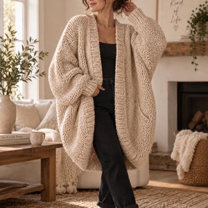 Puede incluir: Un cárdigan de punto grueso y extragrande en tono beige claro, con mangas largas, puños acanalados y una solapa frontal ancha y acanalada. La prenda se lleva sobre una camiseta sin mangas negra y vaqueros gris oscuro, con calcetines acanalados claros.