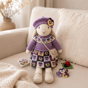 Puede incluir: Una muñeca de conejo tejida que lleva una boina morada con un detalle de pensamiento, un suéter morado con un canesú de pensamientos y una falda con un patrón de cuadros de pensamientos. La muñeca lleva un pequeño bolso cruzado beige.