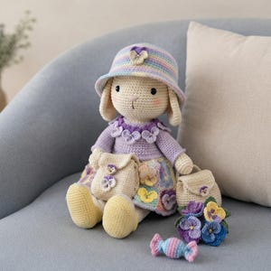 Può includere: Una bambola coniglio all'uncinetto che indossa un cappello a secchiello a righe pastello con un dettaglio a forma di viola, un top color lavanda con colletto floreale e una gonna a motivi. La bambola tiene una borsa beige, con un'altra borsa, un bouquet di viole e una caramella accanto.