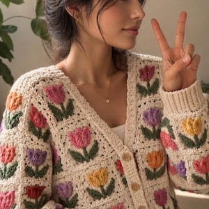 May include: A cream-colored crochet cardigan featuring a repeating pattern of colorful tulip motifs in shades of pink, purple, orange, and yellow with green stems and leaves. The garment has a V-neckline and is fastened with round wooden buttons.