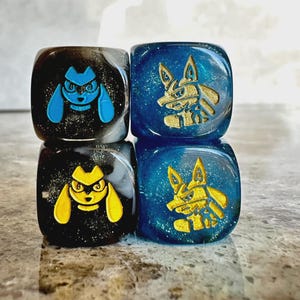 May include: Four square dice with rounded corners. Two black dice feature glitter and engraved character faces in bright blue and yellow. Two blue dice feature glitter and engraved gold character faces. The dice are arranged in a two-by-two grid.