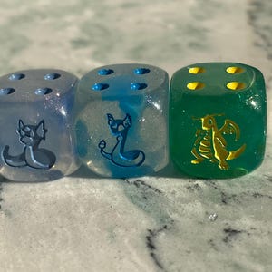 May include: Three six-sided dice with rounded corners. The first is translucent light blue with glitter and a blue dragon-like creature icon. The second is translucent blue and white with a blue creature icon. The third is translucent green with a yellow dragon icon.
