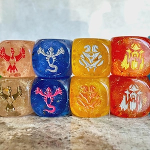 May include: A set of twelve translucent, multi-colored dice arranged in two rows of six. Each die features a unique engraved icon, including stylized creatures and symbols in contrasting colors like green, orange, blue, yellow, and red.