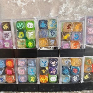 May include: A collection of ten clear plastic display cases, each containing six multi-colored dice. Each die features a unique engraved character icon, including various creatures in shades of green, blue, purple, orange, red, and yellow.