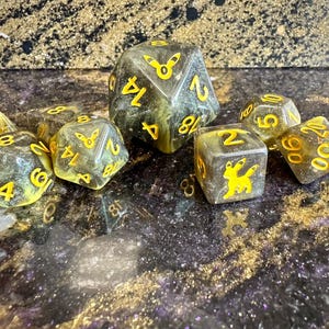 May include: A set of polyhedral dice in a translucent dark olive green with a swirling glitter effect. The dice feature bright yellow numbers and a yellow silhouette of a fox-like creature on the faces. The set includes a d20, d12, d10, d8, d6, and d4.