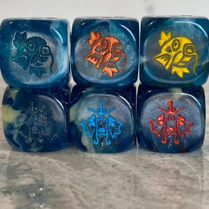 May include: A set of six translucent blue dice with swirling white patterns and glitter. The top row features engraved fish icons in dark blue, orange, and yellow. The bottom row displays engraved dragon-like creature icons in dark blue, light blue, and red.