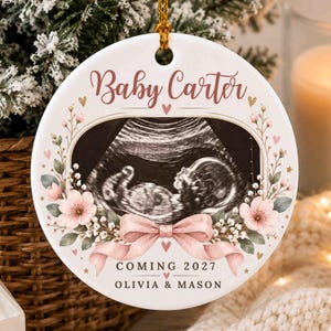 May include: A round white ceramic ornament featuring a central ultrasound image of a fetus. The design includes pink floral accents, a pink ribbon, and the text Baby Carter, COMING 2027, and OLIVIA & MASON. A gold string is attached to the top.