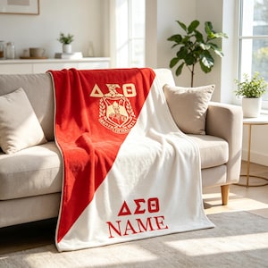Puede incluir: Una manta de felpa con un diseño dividido en diagonal en rojo y blanco. La sección roja muestra un escudo dorado de Delta Sigma Theta y letras griegas. La sección blanca presenta las letras griegas rojas Delta Sigma Theta y la palabra NAME.