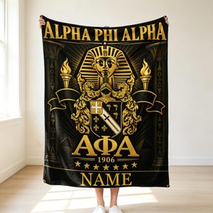 May include: A black and gold blanket featuring the text ALPHA PHI ALPHA at the top, a central pharaoh mask, a heraldic shield with a diagonal stripe, two torches, the Greek letters AΦA, the year 1906, seven stars, and the placeholder text NAME.