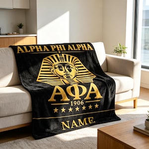 May include: A black fleece throw blanket featuring a gold-coloured graphic of a pharaoh mask, the text ALPHA PHI ALPHA, the Greek letters AΦA, the year 1906, seven stars, and the word NAME printed in a bold, capitalised gold font across the bottom.