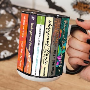 May include: A white ceramic mug with a black handle and interior, featuring a graphic print of six Widespread Panic album spines arranged vertically. The titles include Don't Tell The Band, Ball, Earth To America, Free Somehow, Dirty Side Down, and Street Dogs.