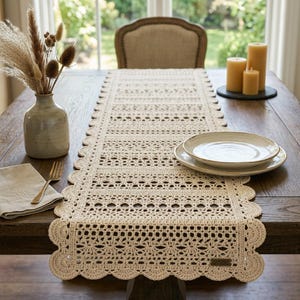 May include: A cream-colored rectangular crochet table runner featuring intricate openwork patterns and a scalloped edge. The runner is displayed on a wooden table alongside a ceramic vase with dried grasses, a linen napkin, and two stacked plates.