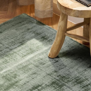 May include: A rectangular area rug featuring a distressed, mottled texture in shades of sage and forest green. The rug is positioned on a wooden floor next to a small, round, light-coloured wooden stool that supports a stack of books and glasses.