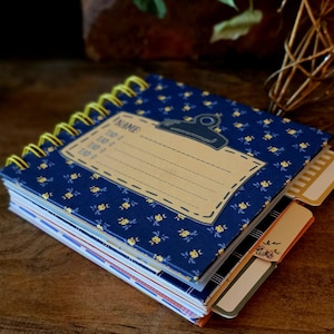 May include: A square spiral-bound notebook with a dark blue cover featuring a small yellow floral pattern. The cover has a tan rectangular label with a clipboard icon and the text NAME, TAD-1, TAD-2, and TAD-3. Several tabbed dividers extend from the side.