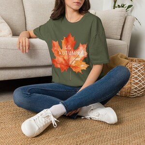 Könnte beinhalten: Ein olivgrünes T-Shirt mit einer großen, aquarellartigen Grafik eines orange-roten Ahornblatts in der Mitte der Brust. Der Text "hello AUTUMN" ist in weißen, serifenlosen Buchstaben quer über die Mitte der Ahornblatt-Illustration gedruckt.
