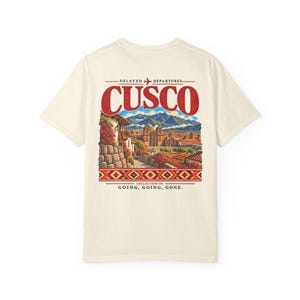 Könnte beinhalten: Ein cremefarbenes T-Shirt mit einem Grafikdruck auf der Rückseite und dem Text "DELAYED DEPARTURES CUSCO COLLECTION 04 GOING, GOING, GONE." Die Illustration zeigt ein farbenfrohes Stadtbild mit Bergen, Steinmauern, Blumen und einer geometrischen Bordüre.
