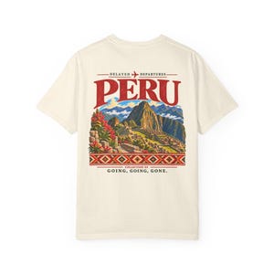Könnte beinhalten: Ein cremefarbenes Kurzarm-T-Shirt mit einem Grafikdruck auf der Rückseite. Das Design enthält den Text DELAYED DEPARTURES, PERU, COLLECTION 04 und GOING, GOING, GONE. Es zeigt eine farbenfrohe Landschaft von Machu Picchu mit Bergen und Blumen.