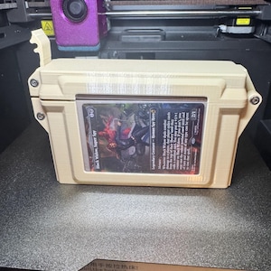 Op de afbeelding: Een beige 3D-geprinte rechthoekige opbergdoos met een doorzichtig venster aan de voorzijde waarin een Black Widow, Super Spy ruilkaart wordt getoond. De doos heeft een scharnierend deksel met een sluitmechanisme en staat voor een 3D-printer.