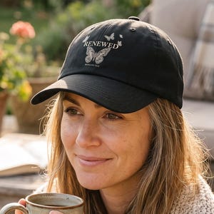 May include: A black baseball cap featuring a graphic of a butterfly and the text RENEWED printed in white on the front panel. The cap has a curved brim and a structured crown, with small butterfly accents surrounding the central text design.