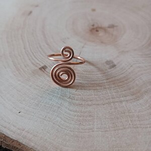Könnte beinhalten: Ein kupferfarbener Drahtring mit zwei verschiedenen Spiralmustern. Die größere Spirale befindet sich unten, während eine kleinere Spirale oben sitzt, verbunden durch ein dünnes, kreisförmiges Drahtband, das die Hauptstruktur des Rings bildet.