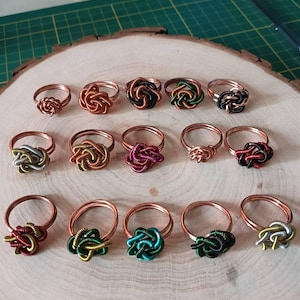 May include: A collection of fourteen wire-wrapped rings arranged in three rows. Each ring features a central knot design crafted from coiled metallic wire in various colors including copper, silver, gold, green, black, red, and teal.
