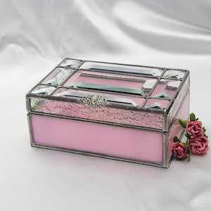 Puede incluir: Una caja rectangular de vidrio manchado rosa con un marco de metal plateado y un cierre decorativo. La tapa presenta detalles de vidrio transparente e iridiscente. Pequeñas rosas rosas se colocan cerca de la caja.