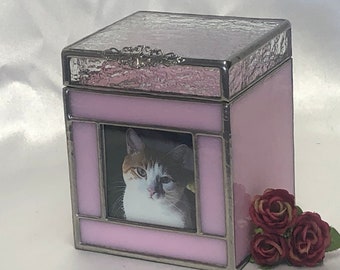 Puede incluir: Una caja de recuerdo de vidrio rosa con un marco plateado y una foto de un gato calico en el interior. La caja tiene una tapa texturizada y está decorada con un diseño plateado.
