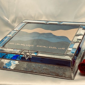 May include: A blue and silver stained glass box with a Star of David on the lid. The box has a blue and white design on the inside and the words "Etz Chaim Hi" are written on the lid.