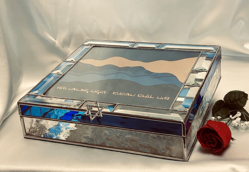 May include: A blue and silver stained glass box with a Star of David on the lid. The box has a blue and white design on the inside and the words "Etz Chaim Hi" are written on the lid.