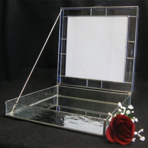 May include: A clear glass box with a hinged lid and a silver chain. The lid has a stained glass frame with a white background. The box is open and the inside is lined with a mirror. A red rose with white flowers is sitting on the box.
